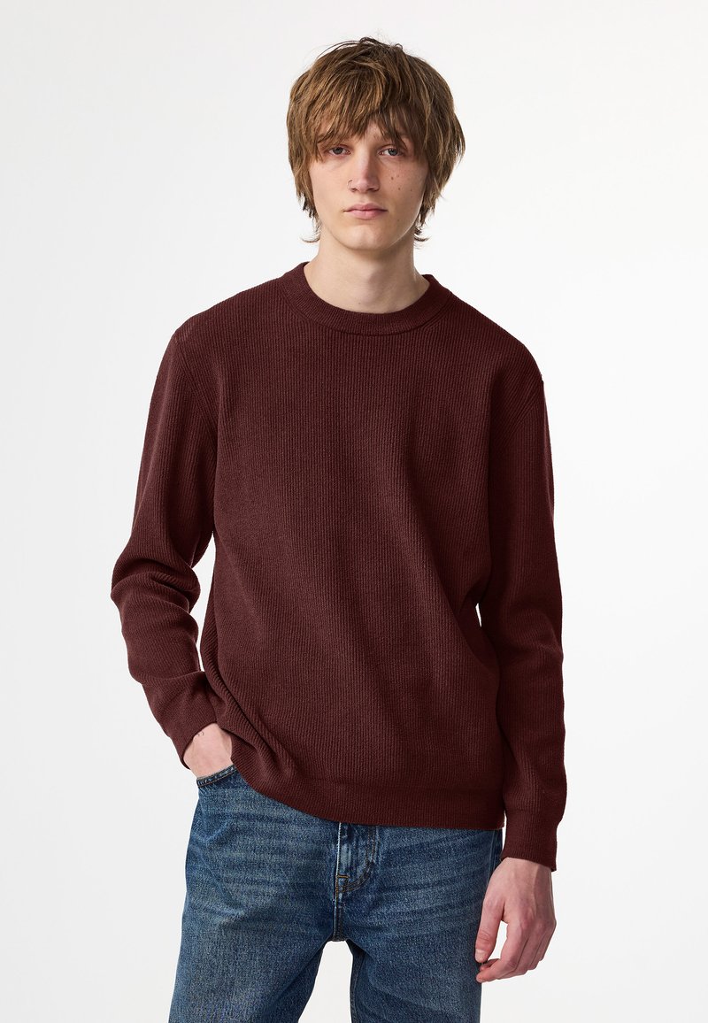 Burgunderfarbener Strickpullover mit strukturiertem Muster, Rundhalsausschnitt und langen Ärmeln. Getragen mit blauen Jeans, Hände in den Taschen.