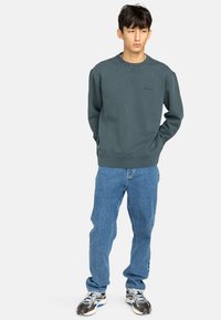 Felpa verde scuro con scollo rotondo, polsini a coste e un sottile logo del marchio. Abbinata a jeans blu chiaro larghi e sneakers.