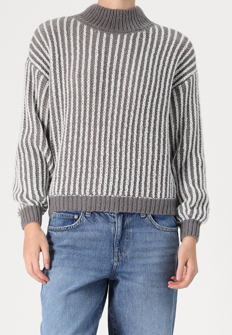 Personne portant un pull en tricot à rayures verticales grises et blanches avec un col, des poignets et un ourlet côtelés, associé à un jean bleu.