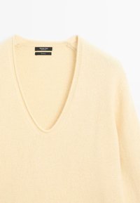 Maglione a V in 100% lana di colore crema morbido. Presenta un tessuto lavorato a maglia e un design pulito e minimalista.