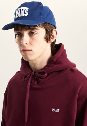 Jonge man met een blauwe Vans-pet en een bordeauxrode Vans-hoodie met trekkoordjes tegen een effen lichte achtergrond.