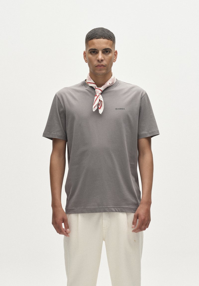 Szürke rövid ujjú póló kerek nyakkivágással és fekete logóval. A nyak körül világospiros-fehér mintás sál van megkötve. Fehér nadrág.