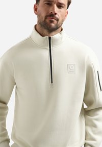 Lichtbeige kwart-zip sweatshirt gemaakt van zacht materiaal, met een zwarte rits, geribde boorden en een vierkante logo patch op de borst.