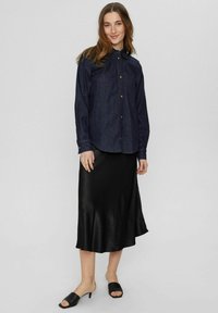 Mörk denimskjorta med knappar och bröstficka, i kombination med en svart satin midi kjol och svarta öppna sandaletter. Släta texturer, avslappnad passform.