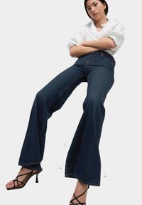 Jeans de mezclilla oscuros de corte acampanado con costuras visibles, cintura alta y una textura de mezclilla suave, combinados con tacones negros con tiras y una camisa blanca.