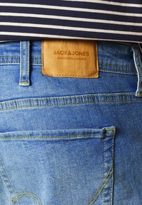 Ljusblå denimjeans med en läderetikett präglad med "JACK & JONES JEANS INTELLIGENCE." Har fickor med gul sömnad.