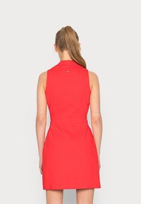 Woman with long ponytail wearing a sleeveless, knee-length red dress viewed from the back against a plain light background.