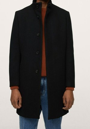 Personne portant un manteau noir boutonné par-dessus un pull couleur rouille et un jean bleu, debout devant un fond clair uni.