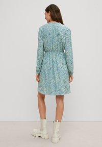 Abito a fantasia floreale nelle tonalità di blu e verde, con maniche lunghe, vita elastica, lunghezza al ginocchio. Indossato con stivali bianchi alla caviglia.