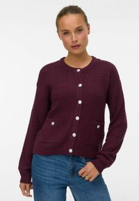 Bordeaux gebreide cardigan met een textuurontwerp, voorzien van een ronde halslijn, zeven knopen en twee voorzakken. Blauwe denim jeans worden gedragen.