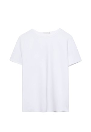 Frau mit schwarzer Lederjacke, weißem Oberteil, Jeansminirock und schwarzen kniehohen Stiefeln lehnt an einer weißen Wand.