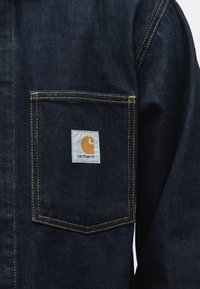 Tmavě modrá džínová bunda s kapsou se žlutým prošitím a bílou čtvercovou nášivkou s logem Carhartt uprostřed kapsy.