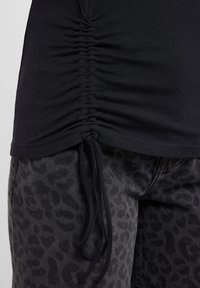 Chemise noire avec côtés froncés et cordons, associée à un jean gris avec un motif léopard subtil. Texture de tissu lisse.