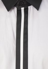 Kraagshirt met een zwarte halslijn en een centrale witte strook, die contrasteert met de witte stof. Gladde textuur, knoopsluiting zichtbaar.