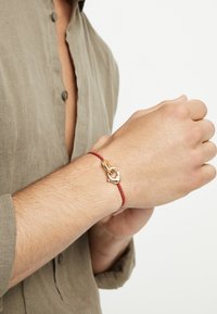 Pulsera trenzada roja con cierre en tono dorado. El diseño presenta un detalle de nudo simple y un acabado liso y brillante en el hardware.
