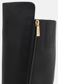 Bota negra de cuero hasta la rodilla con cremallera dorada y acentos texturizados en el costado. Acabado liso con detalle de estrías verticales.