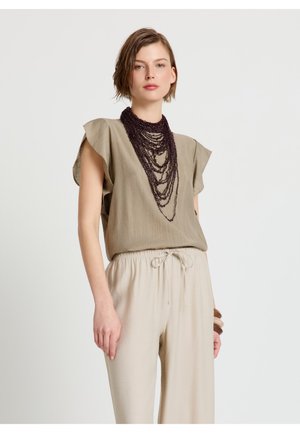 Frau trägt ein hellbraunes Top mit kurzen Ärmeln, cremefarbene Hosen mit Kordelzug, mehrlagige dunkle Perlenkette und braune Armbänder vor weißem Hintergrund.