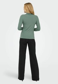 Muntgroene geribde top met lange mouwen en een hoge hals, gecombineerd met zwarte wijde broek. De top heeft een licht viste rand.