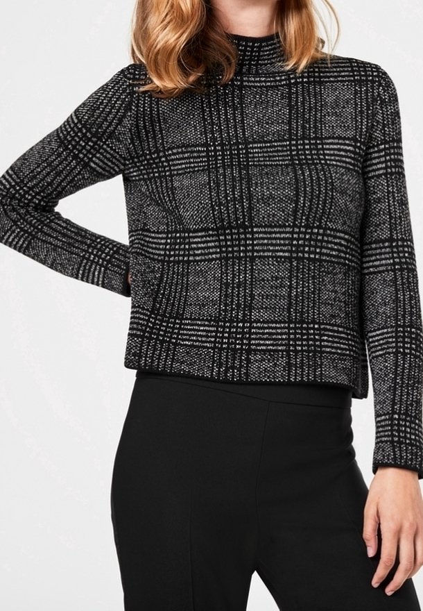 Pull à carreaux noirs et blancs avec un col montant et des manches longues, présentant un tissu texturé et une coupe décontractée. Associé à un pantalon noir.