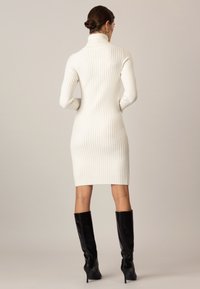 Vit ribbad stickad klänning med hög krage, figursydd design och med längd till mitten av låret. Tillsammans med svarta knähöga stilettkängor.