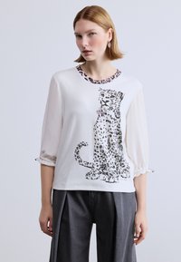 Λευκό μπλουζάκι με λεπτομέρειες από λεοπάρδαλη γύρω από το λαιμό, με γραφικό μιας καθιστής λεοπάρδαλης σε μαύρο και γκρι στο μπροστινό μέρος.