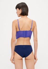 Lilla bralette med justerbare stropper, hægte lukning bagpå, parret med mørkeblå bikiniunderdel, der har en glat tekstur.
