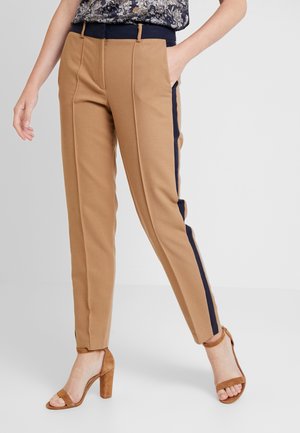 Person trägt beige, schmal zulaufende Hose mit schwarzen Seitenstreifen und braune Sandalen mit Absatz, steht mit einer Hand in der Tasche.