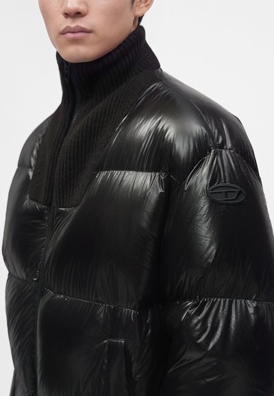 Homme portant une doudoune noire brillante avec un col côtelé haut et un petit patch logo sur le haut de la manche.