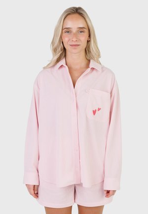 Kobieta o blond włosach, ubrana w jasno różową koszulę na guziki z dwoma małymi czerwonymi sercami na kieszeni piersiowej oraz dopasowane spodenki.