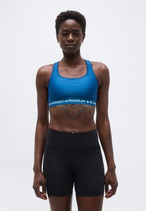 Sportliche Frau mit kurzen lockigen Haaren, die einen blauen Under Armour Sport-BH und schwarze, hoch taillierte Sportshorts trägt, vor weißem Hintergrund stehend.