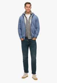 Haut à capuche bleu clair avec cordons de serrage sur une chemise à carreaux, associé à un jean sombre et des baskets beiges. Design simple avec des éléments décontractés.