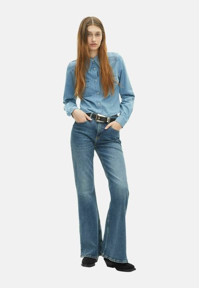 Camicia in denim blu chiaro con colletto a punta e due tasche sul petto, abbinata a jeans blu a campana e stivali neri con tacco. Design semplice.