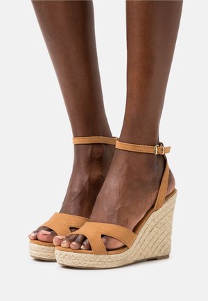 Sandales à talons en toile couleur tan avec une semelle en jute tressé. Elles disposent de deux larges brides à l'avant et d'une bride arrière réglable. Texture douce en daim.