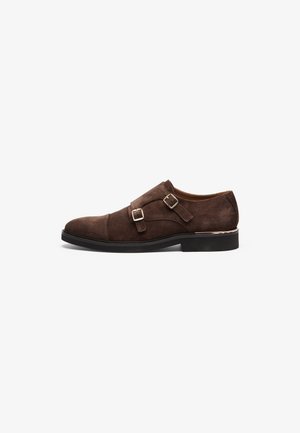 Braune Wildleder-Monk-Strap-Schuhe mit zwei Schnallen, runder Spitze, niedrigem schwarzen Absatz und dezenten Steppdetails auf einem schlichten Hintergrund.