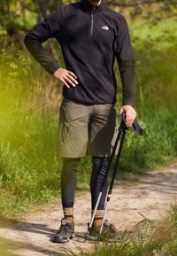 Pullover di pile nero, colletto con zip, pantaloni cargo verdi, leggings neri, scarponi da trekking grigi e bastoni da trekking su un sentiero naturale.