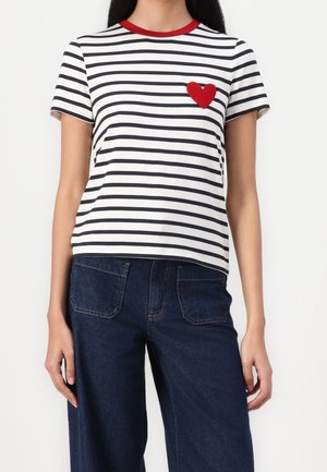 Kvinde iført en kortærmet hvid skjorte med sorte vandrette striber, rød krave og et rødt hjertepatch på brystet, stylet med mørkeblå jeans.