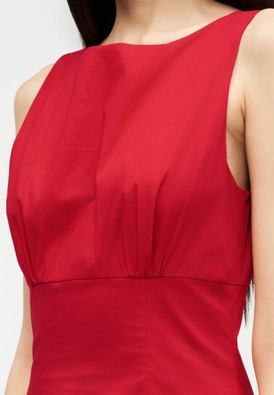 Γυναίκα που φοράει αμάνικο κόκκινο φόρεμα με σούρες στη μέση και λαιμόκοψη σκάφος, εμφανίζοντας το άνω μέρος του κορμού και τον λαιμό της πάνω σε απλό φόντο.