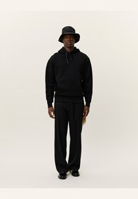 Zwarte katoenen hoodie met trekkoord, frontzak, gecombineerd met zwarte wijde broek en een zwarte bucket hat. Zwarte schoenen maken de outfit compleet.