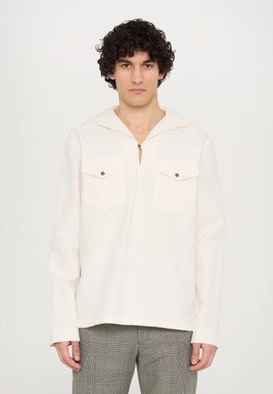 Camicia lunga a maniche con cappuccio di un colore crema chiaro, con placca a bottoni a quarto, due tasche sul petto e un taglio rilassato, abbinata a pantaloni a quadretti.