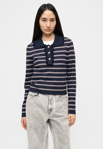 Μπλε ναυτικό τοπ με μακριά μανίκια, βολάν στο γιακά, κούμπωμα με τρία κουμπιά και ρίγες σε λευκό και ροζ αντίθεση· συνδυασμένο με ανοιχτόχρωμο τζιν παντελόνι.