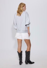 Grå oversized sweatshirt med uppvikt ärm, bärs tillsammans med vita slitna shorts och svarta knähöga cowboystövlar i en studio.