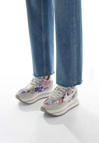 Sneakers with gray suede and shimmering purple/pink floral accents, featuring a thick sole and rounded toe, paired with light blue jeans.
