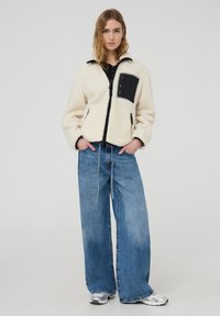 Giacca in pile color crema con dettagli neri, cintura con zip e tasche. Abbinata a jeans larghi di un azzurro chiaro e sneakers argento.