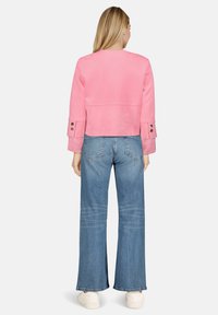 Veste courte en daim rose à manches longues et poignets boutonnés, associée à un jean bleu clair à jambes larges et des baskets blanches.