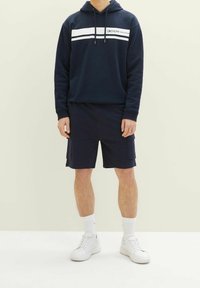 Muž v námořnickém modrém mikině s bílými pruhy, námořnických cargo šortkách, bílých ponožkách a bílých teniskách stojící proti jednobarevnému pozadí.