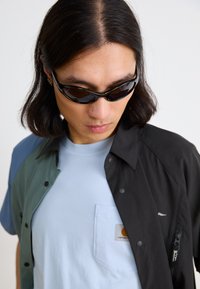 Two-tone shirt in green and black, layered over a light blue t-shirt with a chest pocket. Black sunglasses and a smooth fabric texture.