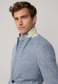 Helleblaue strukturierte Jacke mit einem hohen Kragen und einem cremefarbenen Innenfutter. Vier Knopfdetails und eine einzelne Brusttasche sind sichtbar.
