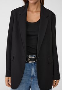 Schwarzer taillierter Blazer mit Reverskragen, ein Knopf und zwei vorderen Taschen, kombiniert mit einem schwarzen gerippten Tanktop und einem schwarzen Gürtel.