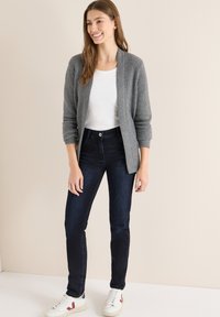 Cárdigan gris acanalado sobre una camiseta blanca, combinado con vaqueros azul oscuro y zapatillas blancas con detalles en rojo. Atuendo simple y casual.