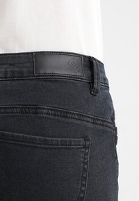 Black denim jeans with a smooth texture, featuring a leather patch on the waistband and a classic five-pocket design.